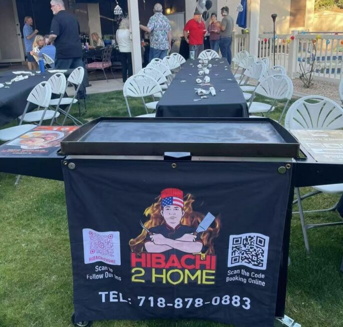Hibachi 2 Home branded portable grill set up on a backyard lawn beside long guest tables for an evening hibachi party at home