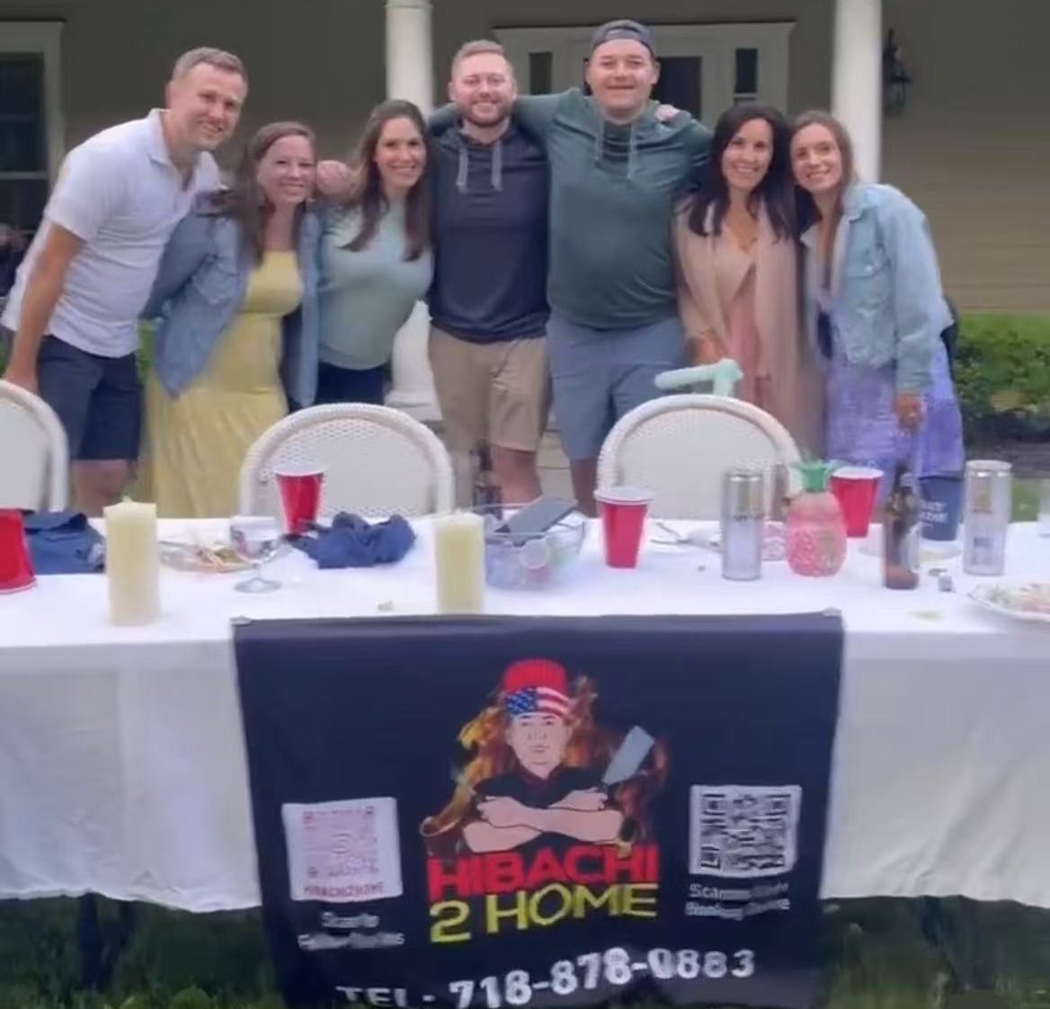 Group of friends posing behind a dinner table with a Hibachi 2 Home banner at a private hibachi party at home
