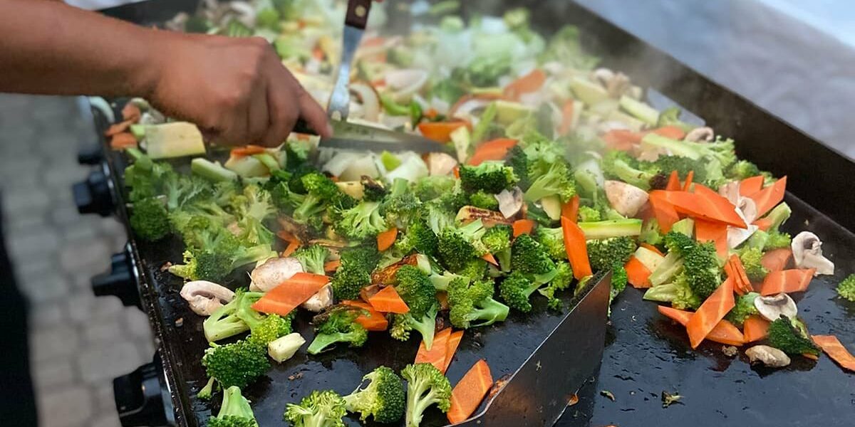 Hibachi chef tossing broccoli, carrots, mushrooms and zucchini on a flat-top grill during a private hibachi dinner
